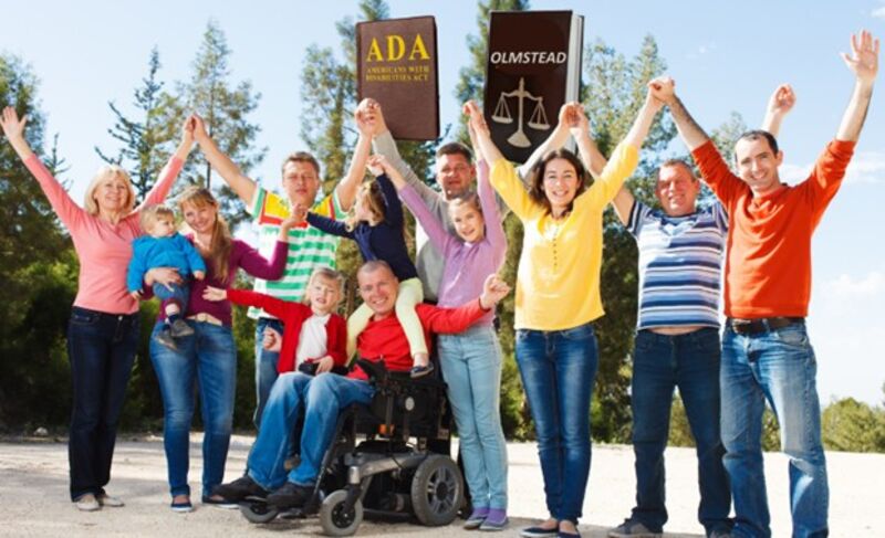 a photo of a group of 12 people, some with visible disabilities, and some without, holding their hands up with law books that say ADA and Olmstead on them, against blue sky and line of trees
