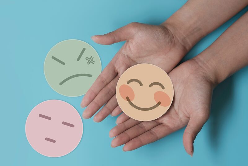 an image of a person's hands holding an emoji smiley face with another emoji angry face and an emoji discontent face on a blue backdrop