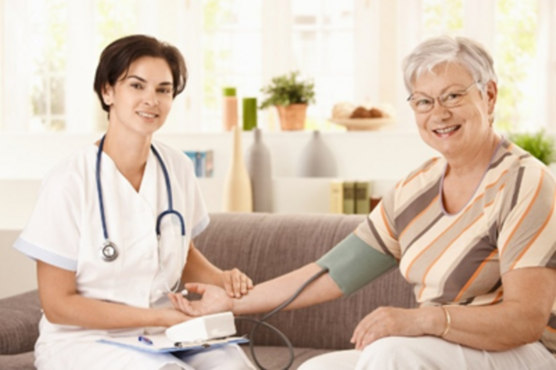 a young brown hair woman who is a nurse is listening to an older woman's blood pressure, both are sitting on the couch smiling at the camera