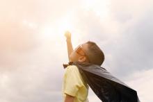 A small boy is dressed as a super hero with a make-shift cape and wide sunglasses and is reaching towards the sky with one hand, intersecting the sun in the background.