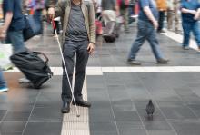 A blind man is navigating through a busy airport using a white cane to follow a textured strip on the ground. 