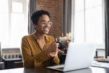 a young black woman wearing her glasses smiling, sitting in front of a laptop, and communicating using sign language in a virtual meeting