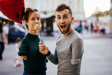 a young straight couple pose for a photo, both with upper limb disabilities. the male holds a heart-shaped lollipop in front of the female's mouth, she is smiling with her eyes and holding a red heart-shaped balloon.
