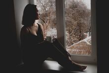 a young woman sitting at her window, staring at the cold weather, with a cup of coffee