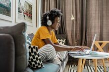 black woman with dark curly hair sitting on a couch, wearing headphones and typing on a laptop placed on a coffee table in front of her