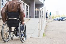 A man wearing a brown leather jacket is about to travel up a ramp using his wheelchair