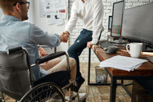 a young white man in a wheelchair is shaking hands with a man that is standing up by his desk