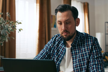 A focused person with a disability is captured in a close-up while working remotely. He sits at home, typing on the keyboard and looking at the monitor with determination.