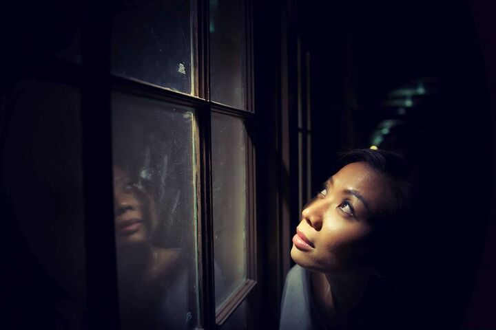 Woman w/ hopeful expression close to a window looking upwards through the window, with a bright light shining down onto her face