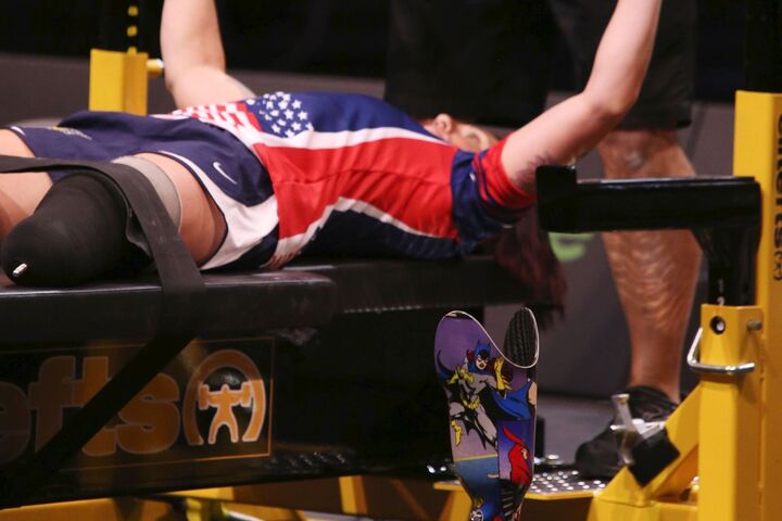 A below the knee amputee is competing in  power lifting bench press, with a spotter ensuring safety and the prosthetic lower limb off the leg, standing by itself in the foreground.