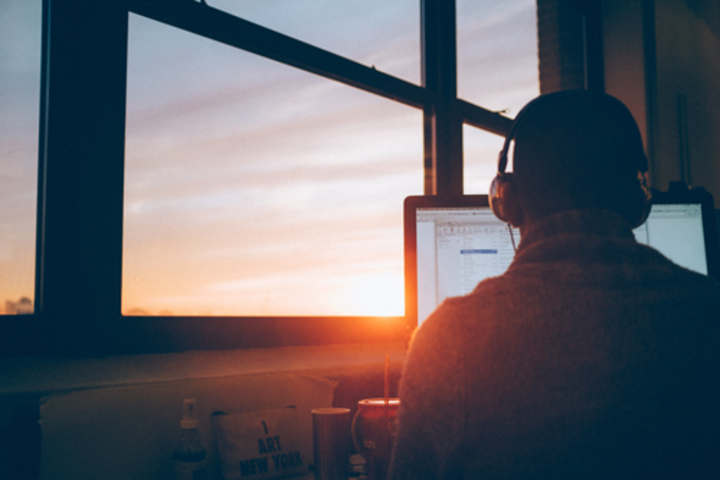 A person sitting at a computer with headphones on overlooking sunset through a window.
