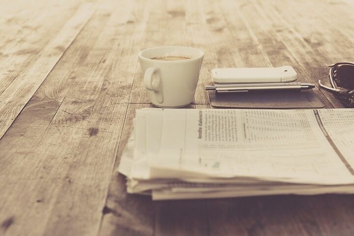 Folded newspaper on a table with a cup of coffee, notepad, pen, and cell phone.