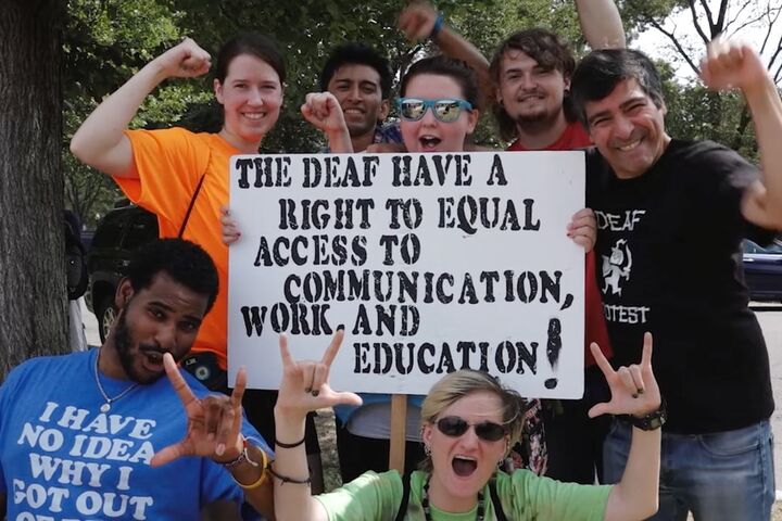 Group of people holding a sign that says THE DEAF HAVE A RIGHT TO EQUAL ACCESS TO COMMUNICATION WORK AND EDUCATION!