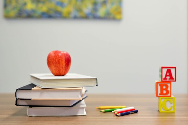 A red apple on top of 3 books next to colored pencils and alphabet blocks.