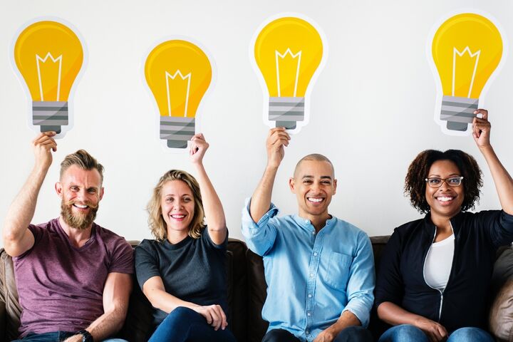 Four smiling people sitting on a couch holding cutouts of light bulbs above their heads to symbolize ideas. 