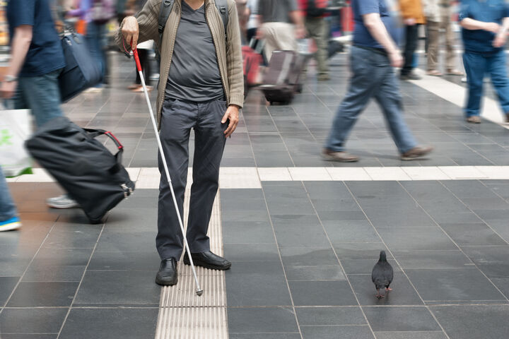 A blind man is navigating through a busy airport using a white cane to follow a textured strip on the ground. 