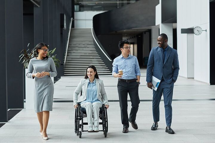 A photograph of a group of diverse coworkers, including a person in a wheelchair, walking and having a conversation in the lobby of a building.