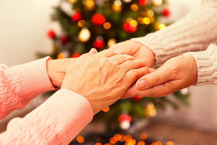 an up-close of an elderly woman's hands, being held by another person with a holiday pine tree in the background