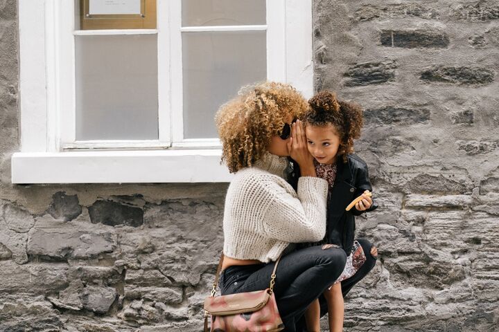 a curly-haired black woman kneels down to her darker curly haired black young daughter to whisper into her ear while standing on a sidewalk of a street