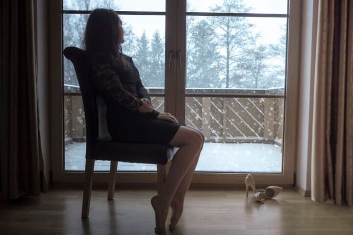 a woman wearing a black dress sitting on a chair staring out the window on a snowy day