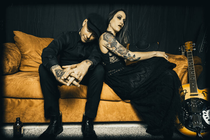 Shelby and Nathen Maxwell dressed in black clothing while sitting on a mustard colored couch.