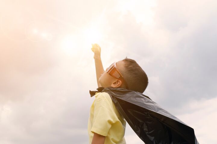 a little boy wears goggles and a superhero cape as he reaches his fist towards a sunlit sky