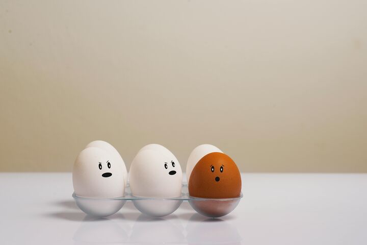 Several white eggs in a carton with faces drawn looking at a brown egg with a face