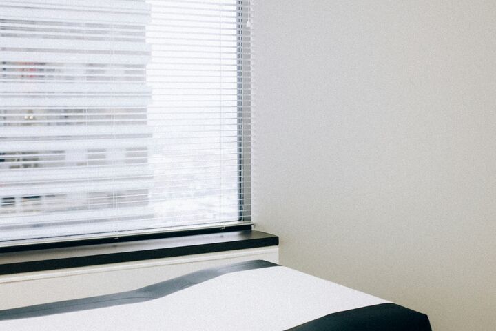 A medical examination table is near a window in a doctor’s office, with paper on top of the table and storage drawers underneath the top of the table.  