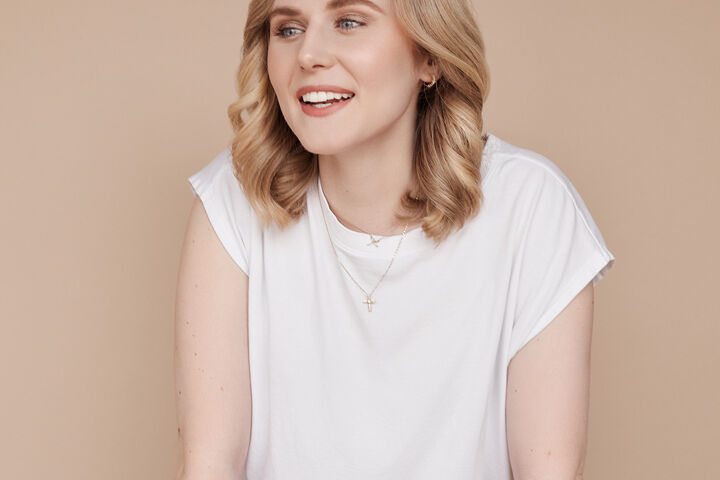 Rebecca Rosenberg, wearing a white tee and brown pants, leans against a brown leather chair.