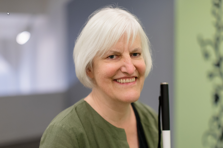 Georgina Kleege wears an olive green cardigan, while smiling and sitting with her cane.