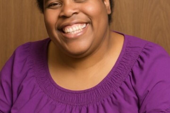 a black woman with short black hair that has a white spot smiling at the camera, wearing a purple shirt on a light brown background