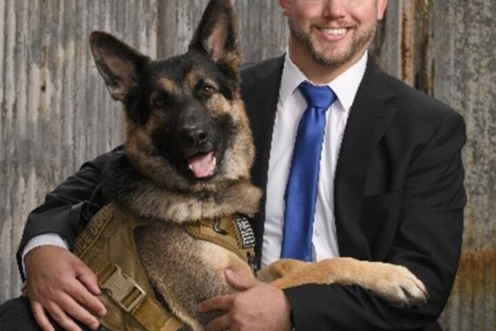 a middle aged bald white man wearing a blue suit sitting and smiling at the camera with his german shepard dog 