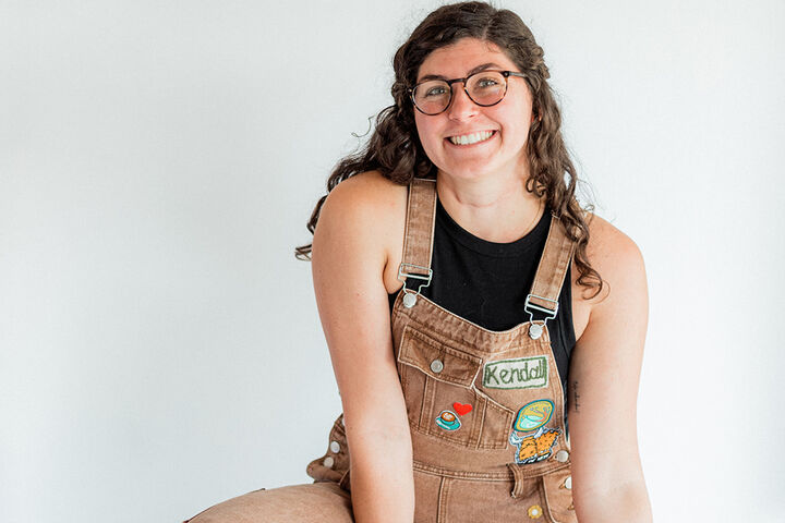 Kendall Peake sits on a wooden stool while wearing brown overalls adorned with patches, a black halter top, and white high top sneakers.