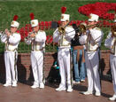 A photo of 5 brass players performing music in the Disneyland Band. The author of this post is center playing trombone in a white marching band uniform. 