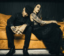 Shelby and Nathen Maxwell dressed in black clothing while sitting on a mustard colored couch.