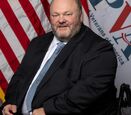 Tom Wheaton dressed in black suit with the U.S.A and PVA flags in the background.