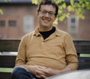 Andrew Leland wears a mustard yellow polo shirt while sitting on a park bench.