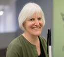 Georgina Kleege wears an olive green cardigan, while smiling and sitting with her cane.