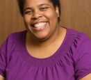 a black woman with short black hair that has a white spot smiling at the camera, wearing a purple shirt on a light brown background