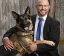 a middle aged bald white man wearing a blue suit sitting and smiling at the camera with his german shepard dog 