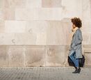 a side profile of a woman wearing a long coat walking on a sidewalk next to the face of a large building