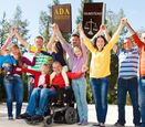 a photo of a group of 12 people, some with visible disabilities, and some without, holding their hands up with law books that say ADA and Olmstead on them, against blue sky and line of trees