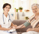 a young brown hair woman who is a nurse is listening to an older woman's blood pressure, both are sitting on the couch smiling at the camera