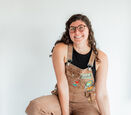 Kendall Peake sits on a wooden stool while wearing brown overalls adorned with patches, a black halter top, and white high top sneakers.